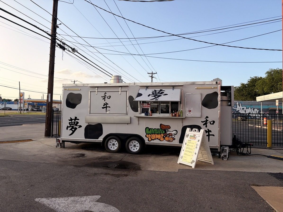 A food truck parked on a corner lot at dusk. Power lines cross the whole sky, a utility pole stands at the left, a traffic cone and cables sit on the ground, and distant cars and a gas station sign clutter the background.