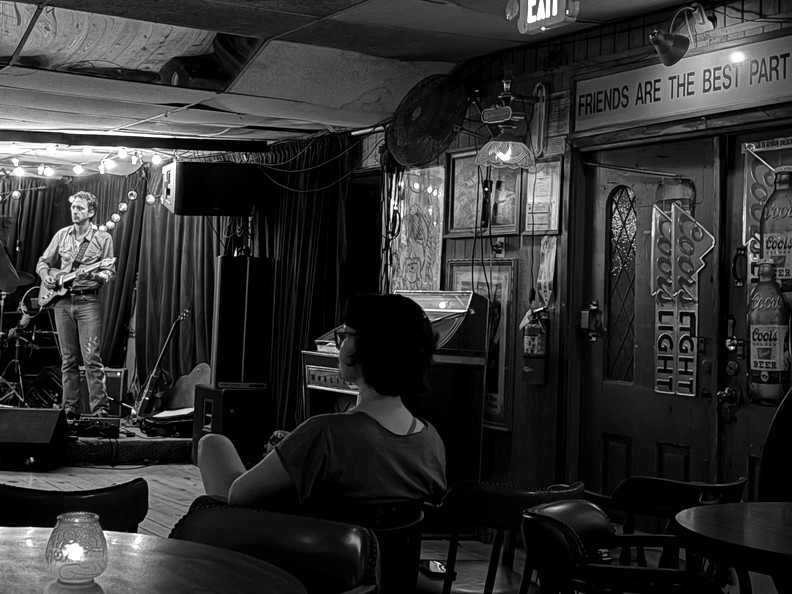 Black-and-white crop of the same frame: the woman at the table watching the guitarist.